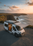 A beautifully finished long-wheelbase camper van parked on a coastal layby overlooking rugged Irish cliffs and the Atlantic Ocean. The van features a clean white exterior with subtle graphite accents, roof rack with solar panels, and an extended awning partially deployed. Inside, warm wooden cabinetry and soft LED strip lighting are visible through the open side door. Captured in photographic realism during golden hour, with low sun casting soft highlights along the van’s bodywork and gentle shadows on the gravel. Shot at eye level with a wide-angle lens, using the rule of thirds to balance van and landscape, creating a professional, aspirational yet practical mood that communicates road-ready freedom and quality craftsmanship.